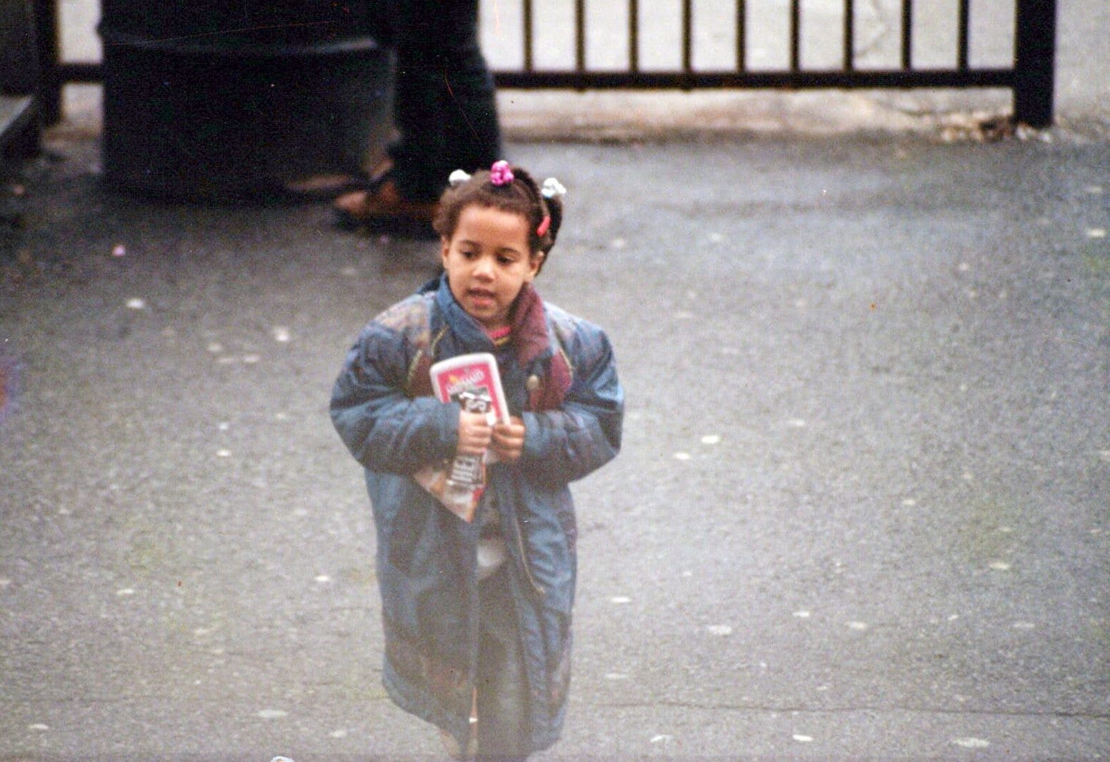 Me as a kid, in an oversized blue puffer coat, walking across a schoolyard holding a handheld game with both hands.