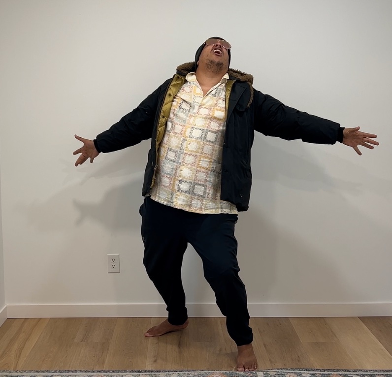 A still from the TrashMan shoot: CJ in a fur-hooded parka and sunglasses, head thrown back and arms flung wide, mid taking-damage pose.