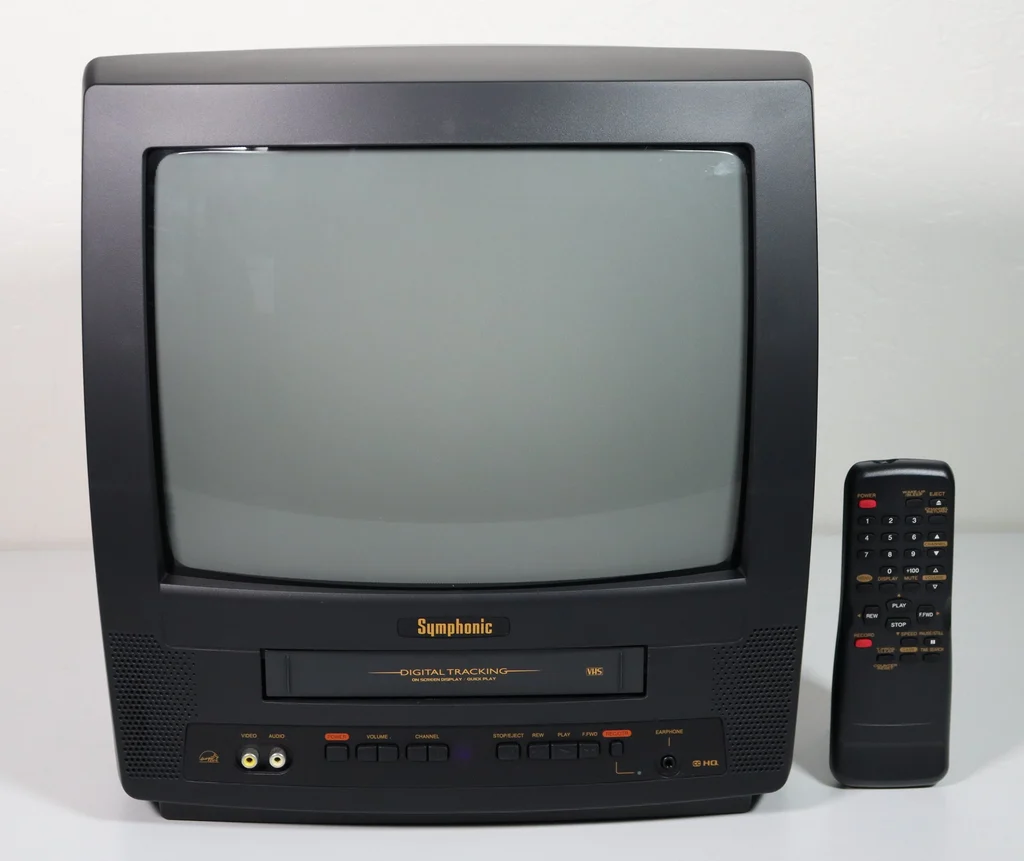 A black Symphonic CRT TV with a built-in VHS player below the screen, its remote propped up beside it.