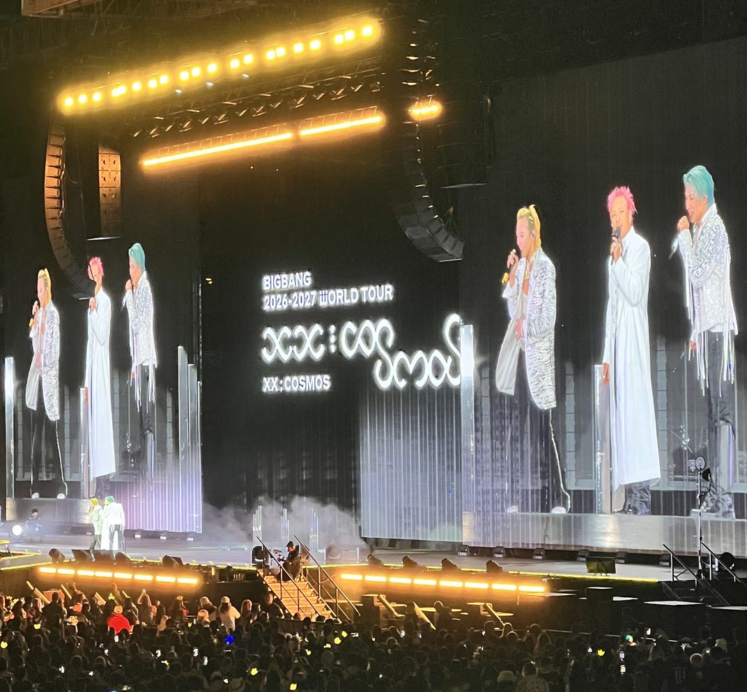BIGBANG on stage in Oakland: G-Dragon, Taeyang and Daesung on the giant screens above a crowd of glowing yellow crown light sticks, the XX: COSMOS tour logo between them.