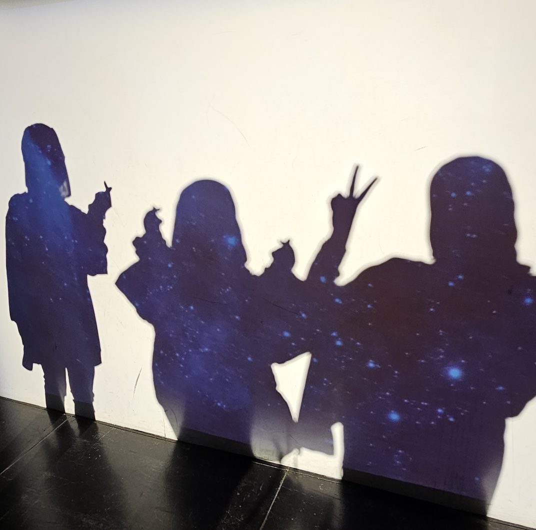 Three shadows cast on a white wall and filled in with a starry galaxy projection: one waving, one holding up a light stick, one throwing a peace sign.