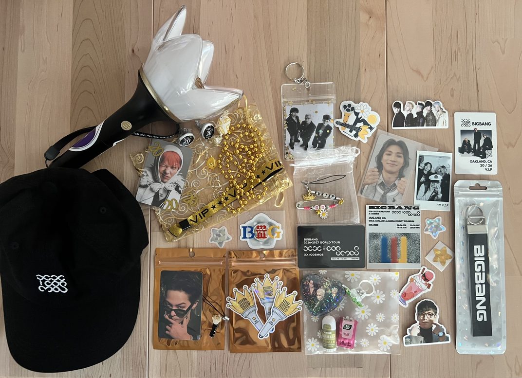 A flat lay of BIGBANG swag on a wood floor: a crown light stick, a black cap, gold beads, a VIP wristband, photocards, stickers, a keychain strap, a phone charm and an Oakland VIP ticket card.