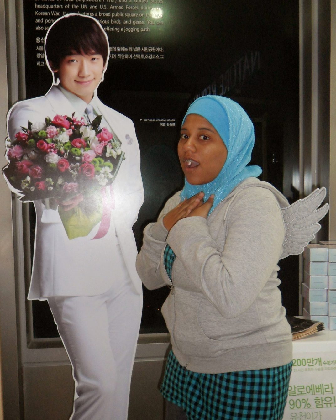 Me in 2011, in a blue hijab and grey hoodie, hand on my chest and mouth open in mock surprise, next to a life-size cardboard cutout of Rain in a white suit holding a bouquet.