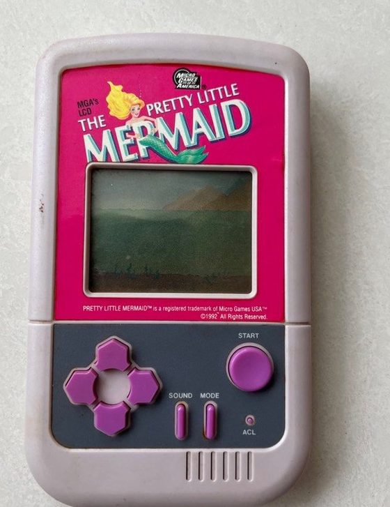 The Pretty Little Mermaid LCD handheld: a grey plastic unit with a hot-pink faceplate, a small green LCD screen, and a purple D-pad and Start button.